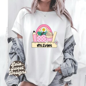 May include: White t-shirt with a teacher-themed design. The design includes a rainbow, pencil, apple, coffee cup, and ruler. The text "Ms. Evans" is printed on a ruler. The shirt is from Lola Sweet Designs.