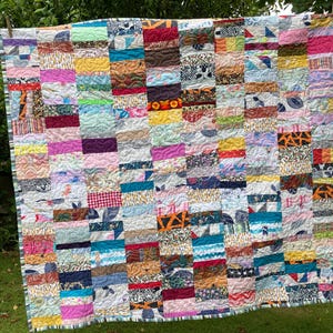 May include: A rectangular patchwork quilt composed of numerous small, horizontal fabric rectangles in a wide variety of colours and patterns, including pink, yellow, blue, orange, and black and white prints, featuring visible wavy machine quilting.