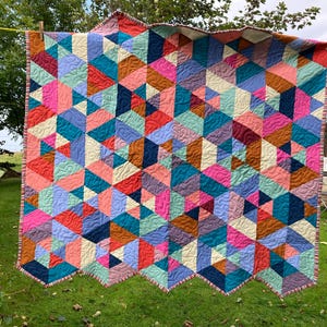 May include: A quilt featuring a geometric pattern of triangles and hexagons in a vibrant palette of pink, teal, navy blue, lavender, coral, and cream. The fabric pieces are joined with visible stitching and finished with a striped fabric border.