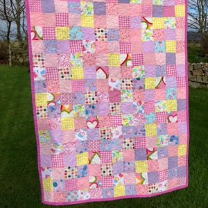 May include: A colourful patchwork quilt with a pink border, hanging on a washing line. The quilt is made up of squares in shades of pink, purple, and yellow, with patterns like hearts, polka dots, and gingham. The quilt is displayed outside on a sunny day.