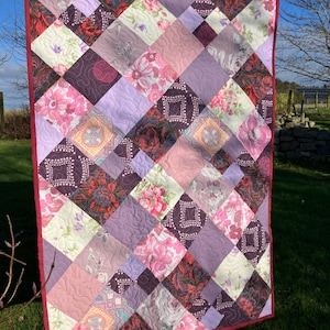 May include: A quilt with a diamond pattern made from various floral fabrics in shades of pink, purple, and red. The quilt is hanging on a washing line outdoors.