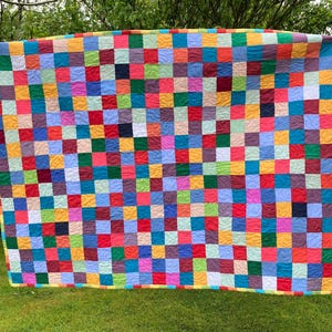 May include: A rectangular quilt featuring a grid of small, multi-coloured fabric squares in shades of red, blue, yellow, green, and purple. The surface displays a consistent swirling quilted texture across the entire piece, finished with a border.