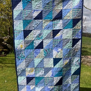 May include: A handcrafted quilt with a patchwork design, showcasing a variety of blue, teal, and grey hues. The quilt is displayed outdoors, with a combination of patterned and plain fabric squares and triangles. It has a dark blue border.