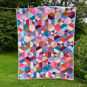 May include: A rectangular quilt featuring a geometric pattern of multicoloured triangles and hexagons in shades of pink, blue, purple, orange, and cream. The fabric surface displays a continuous swirling quilted texture and a light blue striped border.
