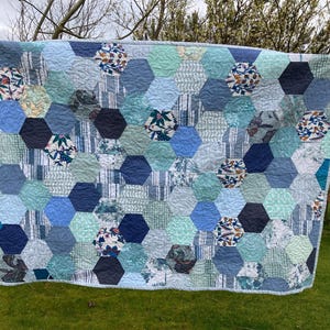 May include: A patchwork quilt with a hexagonal design in various shades of blue and grey. The quilt incorporates floral and striped fabric patterns. It is displayed outdoors, hanging on a washing line, highlighting its handmade quality and design.