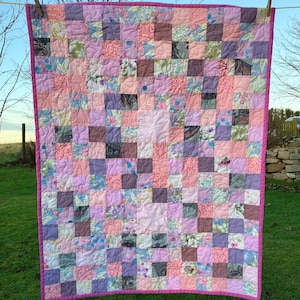 May include: A patchwork quilt with a pink border, hanging on a washing line. The quilt is made from squares of floral and patterned fabrics in pinks, purples, and peach, arranged in a grid. The quilt is displayed outdoors, with a green lawn in the background.