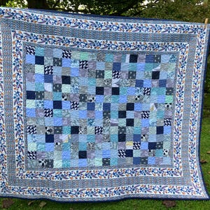 May include: A square patchwork quilt featuring a grid of blue, light blue, and white fabric squares with various patterns. The quilt is framed by a wide border decorated with a repeating blue and orange floral vine motif on a white background.
