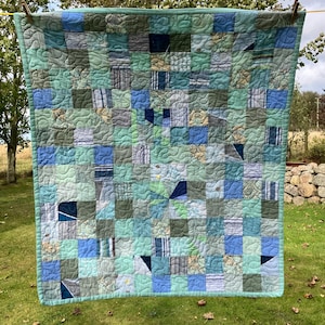 May include: A patchwork quilt in shades of blue, green, and grey, hanging on a washing line. The quilt is made up of numerous squares with different patterns and textures. The overall design is a mix of cool tones, creating a calming effect.