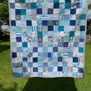 May include: A patchwork quilt featuring a grid of square fabric blocks in various shades of blue, including navy, teal, sky blue, and white. The quilt displays a mix of solid colours and patterned prints, finished with a grey striped border.