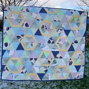 May include: A patchwork quilt with a geometric design. The quilt features triangles and hexagons in shades of blue, green, and white, with yellow accents. The quilt is bordered with a dark blue fabric. The quilt is hanging outside.