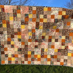 May include: A handmade patchwork quilt in shades of brown, orange, yellow, and pink. The quilt is square-shaped with a black and white patterned border. The quilt is hanging outside on a washing line. The quilt is made of various fabrics.
