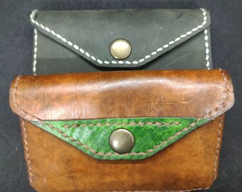 May include: Two leather wallets, one brown with a green flap and one black with a white flap. Both wallets have a metal button closure.