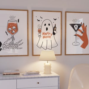 May include: Three framed Halloween art prints. One shows a skeleton hand holding a wine glass with skulls. Another features a ghost with the text "Happy Hour". The third depicts a hand holding a martini glass with a skull.