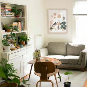 May include: A cozy living room scene with a gray sofa, wooden furniture, and a framed art print. The room is filled with potted plants and a bookshelf. The art print depicts a cat at a table with plants.