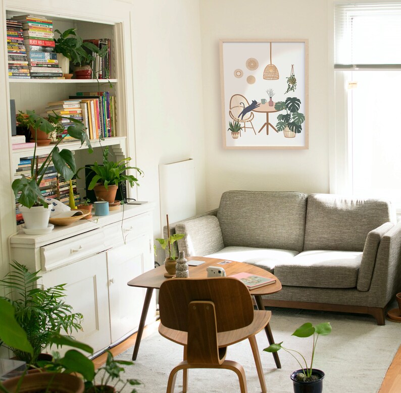 May include: A cozy living room scene with a gray sofa, wooden furniture, and a framed art print. The room is filled with potted plants and a bookshelf. The art print depicts a cat at a table with plants.