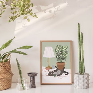 May include: A framed art print featuring a cat lounging near a potted plant and a lamp. The artwork has a light green background and a wooden frame. A cactus in a concrete pot and other plants are also visible.