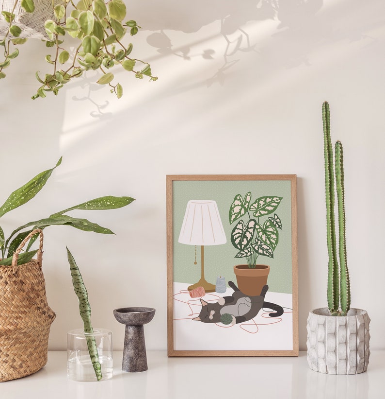 May include: A framed art print featuring a cat lounging near a potted plant and a lamp. The artwork has a light green background and a wooden frame. A cactus in a concrete pot and other plants are also visible.