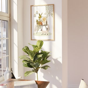 May include: A framed art print featuring a whimsical illustration of a table adorned with potted plants, a bottle, and a cat. The artwork is in a light-filled room with a large window and a green leafy plant in the foreground. The frame is a light wood color.
