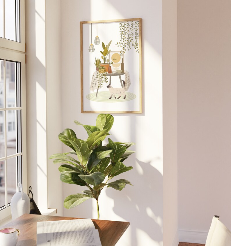 May include: A framed art print featuring a whimsical illustration of a table adorned with potted plants, a bottle, and a cat. The artwork is in a light-filled room with a large window and a green leafy plant in the foreground. The frame is a light wood color.