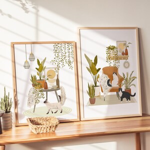May include: Two framed art prints featuring interior scenes with cats and plants. One print shows a cat near a table with plants and hanging lights. The other print depicts a cat in an armchair with plants and decorative items. Both prints have wooden frames.