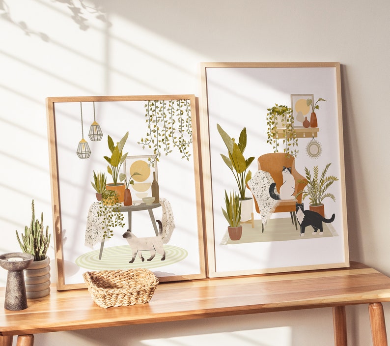 May include: Two framed art prints featuring interior scenes with cats and plants. One print shows a cat near a table with plants and hanging lights. The other print depicts a cat in an armchair with plants and decorative items. Both prints have wooden frames.