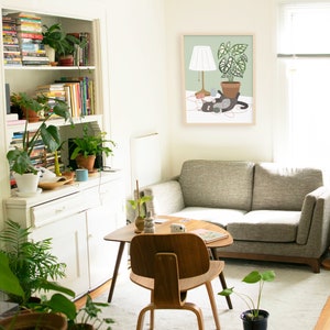 May include: A cozy living room scene with a gray sofa, wooden chair, and a small coffee table. A framed print depicts a cat with a lamp and potted plant. Bookshelves and various potted plants add to the room's inviting atmosphere.