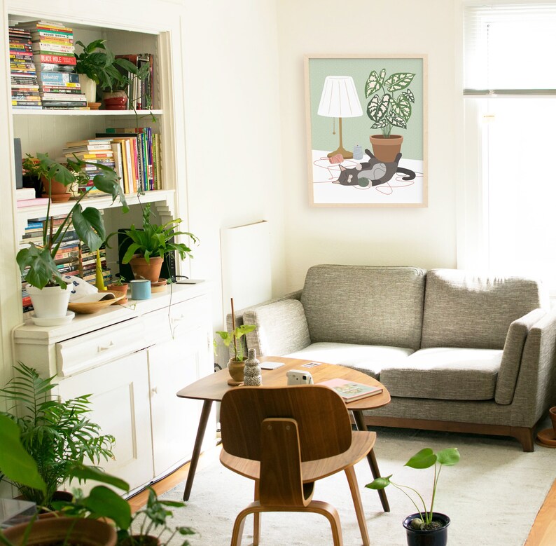 May include: A cozy living room scene with a gray sofa, wooden chair, and a small coffee table. A framed print depicts a cat with a lamp and potted plant. Bookshelves and various potted plants add to the room's inviting atmosphere.