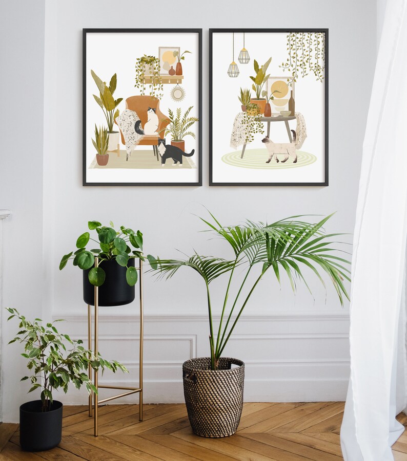 May include: Two framed art prints depict interior scenes with cats, plants, and furniture. One print shows a cat on a chair, the other a cat near a table. The image also includes potted plants and a gold plant stand.