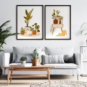 May include: Two framed art prints featuring illustrations of plants and cats. One print shows a cat among potted plants. The other print depicts a cat stretching near a table with plants. The prints are displayed above a white sofa with decorative pillows.