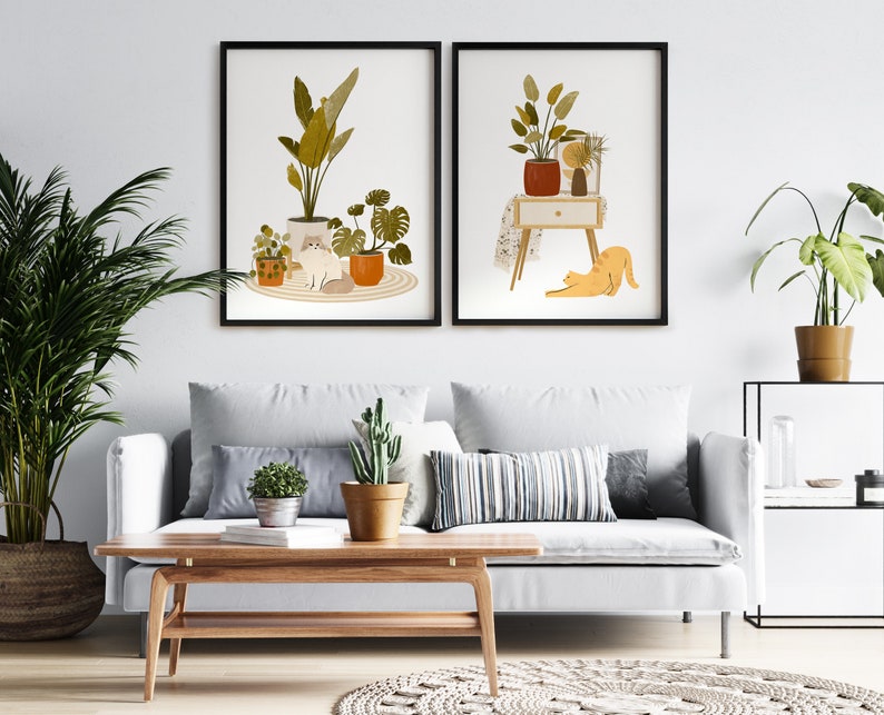 May include: Two framed art prints featuring illustrations of plants and cats. One print shows a cat among potted plants. The other print depicts a cat stretching near a table with plants. The prints are displayed above a white sofa with decorative pillows.