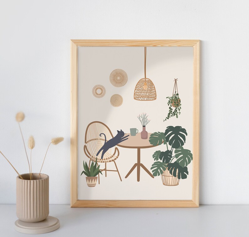 May include: A framed art print featuring a whimsical interior scene. A black cat leaps over a table set with a mug and vase. The scene includes a woven chair, hanging plants, and a woven pendant light. The color palette is neutral with green and brown accents.
