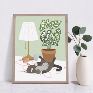 May include: A framed art print featuring a gray cat playing with yarn. A potted plant, a lamp, and a vase with greenery are also depicted. The background is a light green color, and the frame is a light brown color.