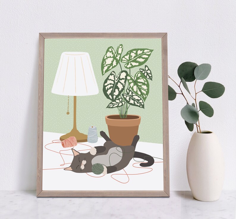 May include: A framed art print featuring a gray cat playing with yarn. A potted plant, a lamp, and a vase with greenery are also depicted. The background is a light green color, and the frame is a light brown color.