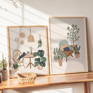 May include: Two framed art prints featuring interior scenes with a bohemian aesthetic. One print depicts a cat jumping towards a table with plants, the other shows a cat resting on a chair surrounded by potted plants. Both prints have wooden frames.