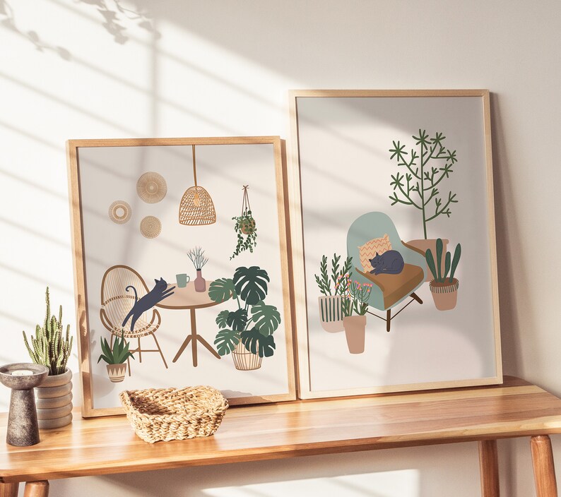 May include: Two framed art prints featuring interior scenes with a bohemian aesthetic. One print depicts a cat jumping towards a table with plants, the other shows a cat resting on a chair surrounded by potted plants. Both prints have wooden frames.