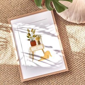 May include: A framed art print featuring a minimalist illustration of a small table with potted plants and a stretching orange cat. The artwork is in a light wood frame, set against a neutral background.