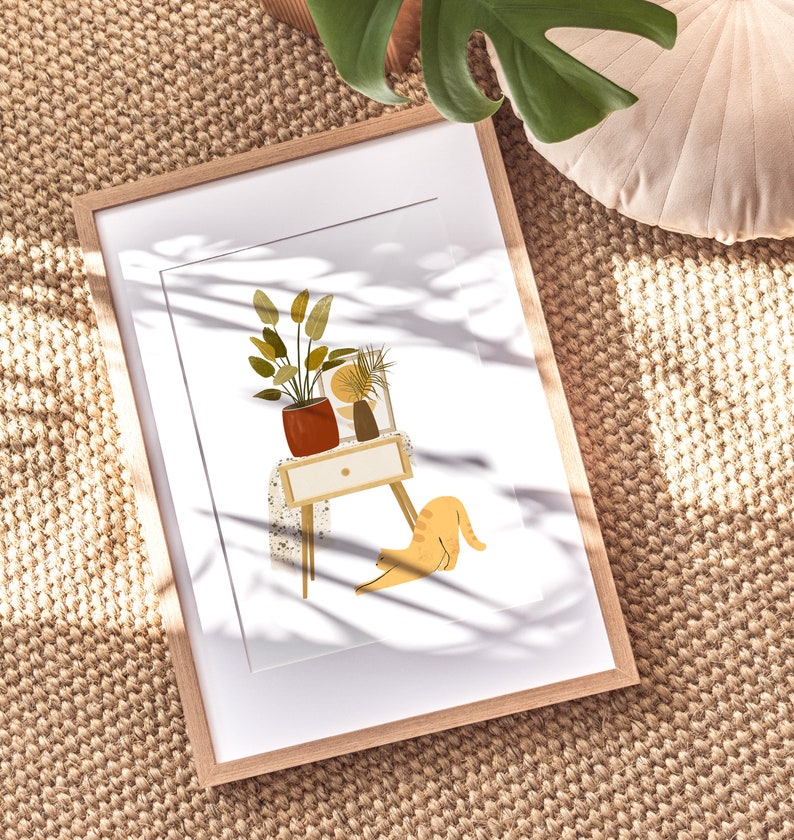 May include: A framed art print featuring a minimalist illustration of a small table with potted plants and a stretching orange cat. The artwork is in a light wood frame, set against a neutral background.