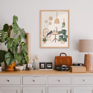 May include: A framed art print featuring a cat leaping towards a table with plants and a hanging light. The print is in a light wood frame and is displayed on a wooden cabinet with a lamp, plants, and a leather suitcase. The color palette is neutral with green and brown accents.