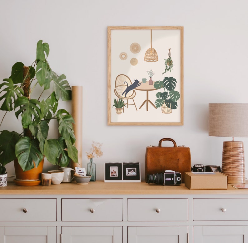May include: A framed art print featuring a cat leaping towards a table with plants and a hanging light. The print is in a light wood frame and is displayed on a wooden cabinet with a lamp, plants, and a leather suitcase. The color palette is neutral with green and brown accents.