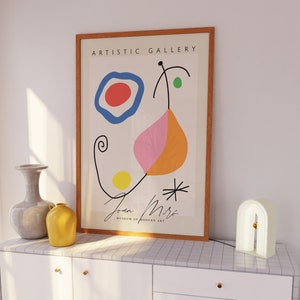 May include: A framed print of a Joan Miró abstract artwork with the text "ARTISTIC GALLERY" at the top. The artwork features colorful shapes and lines. A gray vase, a yellow vase, and a white arched lamp are on a white cabinet.