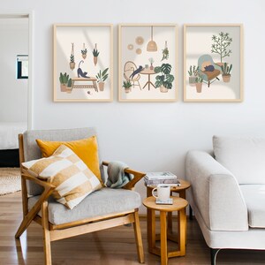 May include: Three framed art prints featuring illustrations of cats in cozy indoor settings with plants. The prints have a neutral color palette with light brown frames. The artwork depicts cats stretching, lounging, and interacting with plants and furniture.