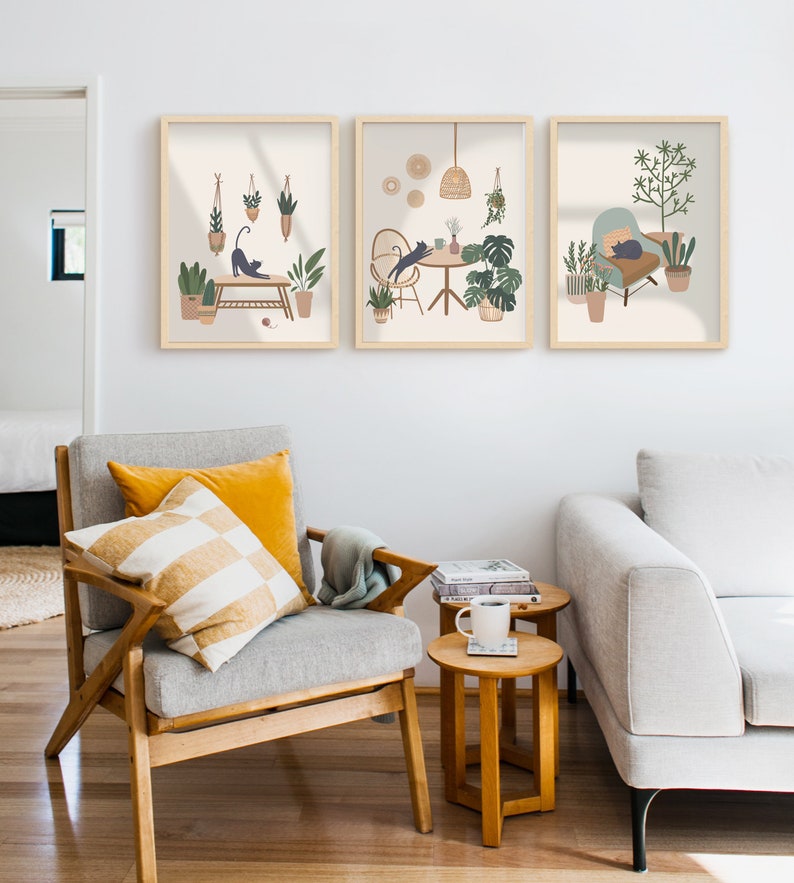 May include: Three framed art prints featuring illustrations of cats in cozy indoor settings with plants. The prints have a neutral color palette with light brown frames. The artwork depicts cats stretching, lounging, and interacting with plants and furniture.