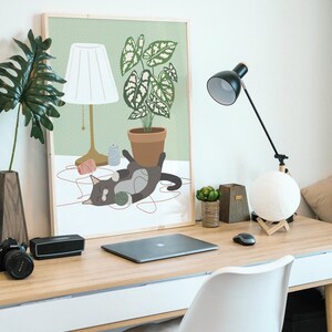 May include: A framed art print depicts a cat playing with yarn near a potted plant and a lamp. A black desk lamp, a moon-shaped light, and a laptop are also on the wooden desk. A camera and a speaker are on the left side of the desk.