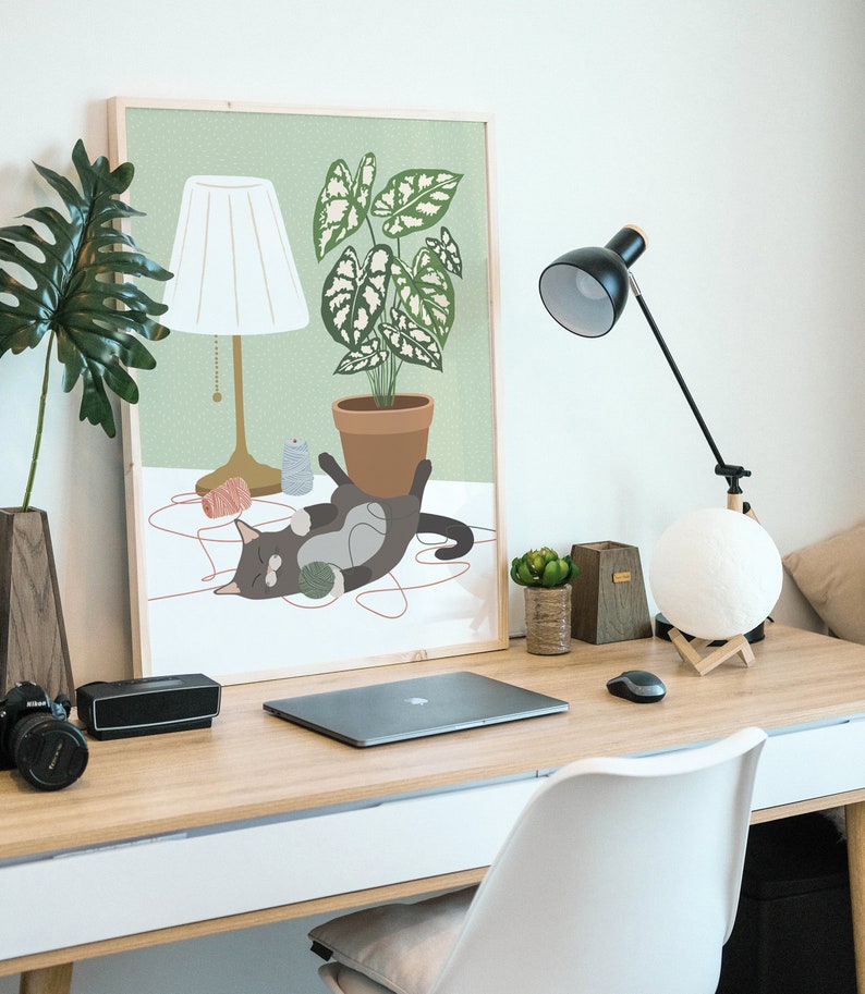 May include: A framed art print depicts a cat playing with yarn near a potted plant and a lamp. A black desk lamp, a moon-shaped light, and a laptop are also on the wooden desk. A camera and a speaker are on the left side of the desk.