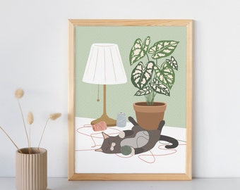 May include: A framed art print featuring a playful cat with a ball of yarn, a potted plant, and a lamp. The illustration has a light green background with a white table and a wooden frame. The cat is gray and white.
