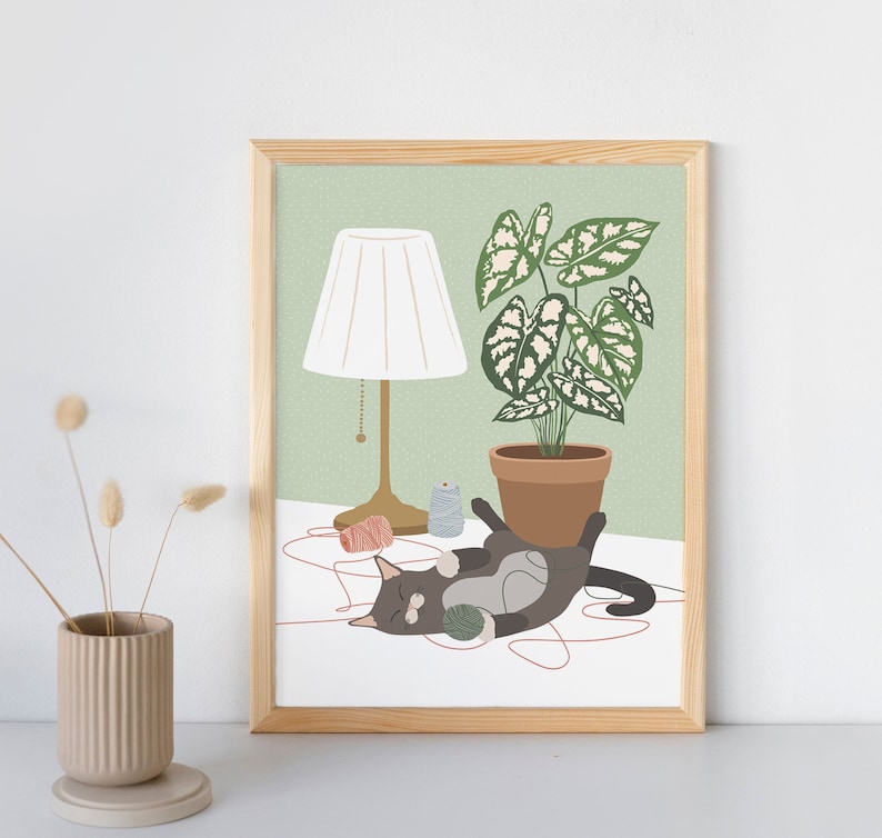 May include: A framed art print featuring a playful cat with a ball of yarn, a potted plant, and a lamp. The illustration has a light green background with a white table and a wooden frame. The cat is gray and white.