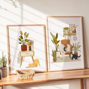 May include: Two framed art prints featuring illustrations of cozy interior scenes with cats and plants. One print shows a cat stretching near a side table with a plant. The other print depicts cats on a chair surrounded by plants.