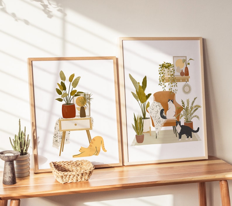 May include: Two framed art prints featuring illustrations of cozy interior scenes with cats and plants. One print shows a cat stretching near a side table with a plant. The other print depicts cats on a chair surrounded by plants.