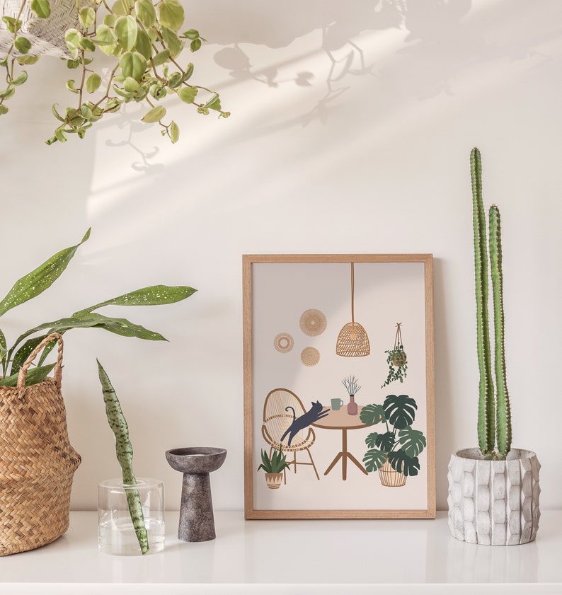 May include: A framed print with a cat leaping over a table, surrounded by plants and a woven lamp. The print is in a light wood frame. A tall cactus in a concrete pot is on the right. Other plants are in woven baskets and glass vases.