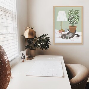May include: A framed art print featuring a playful cat with yarn, a potted plant, and a lamp. The artwork has a light green background and a wooden frame. A gold desk lamp and a small potted plant are also visible.